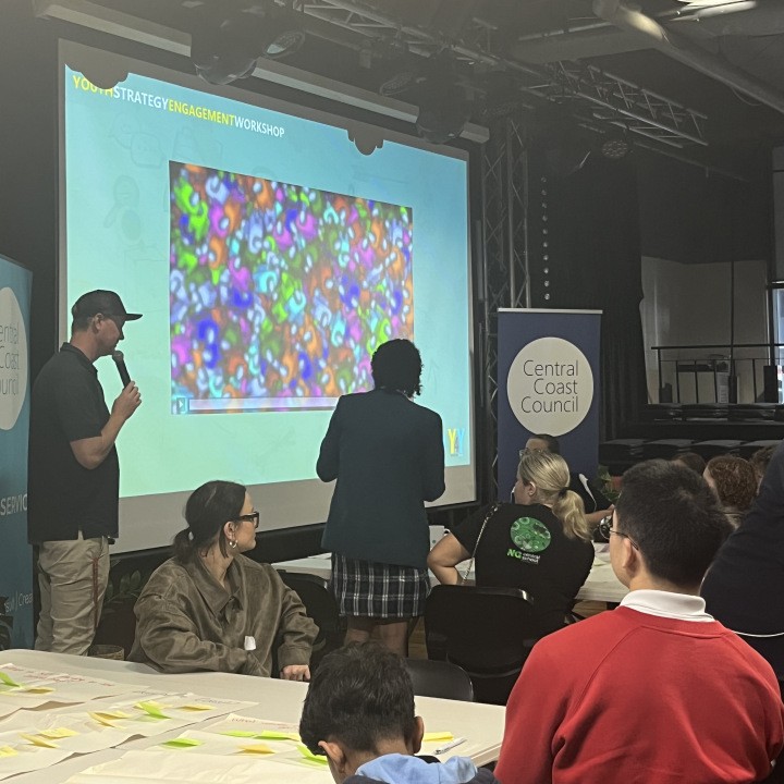 A man standing at a powrpoint slide with a microphone, a students standing up and other students sitting at a table. 
