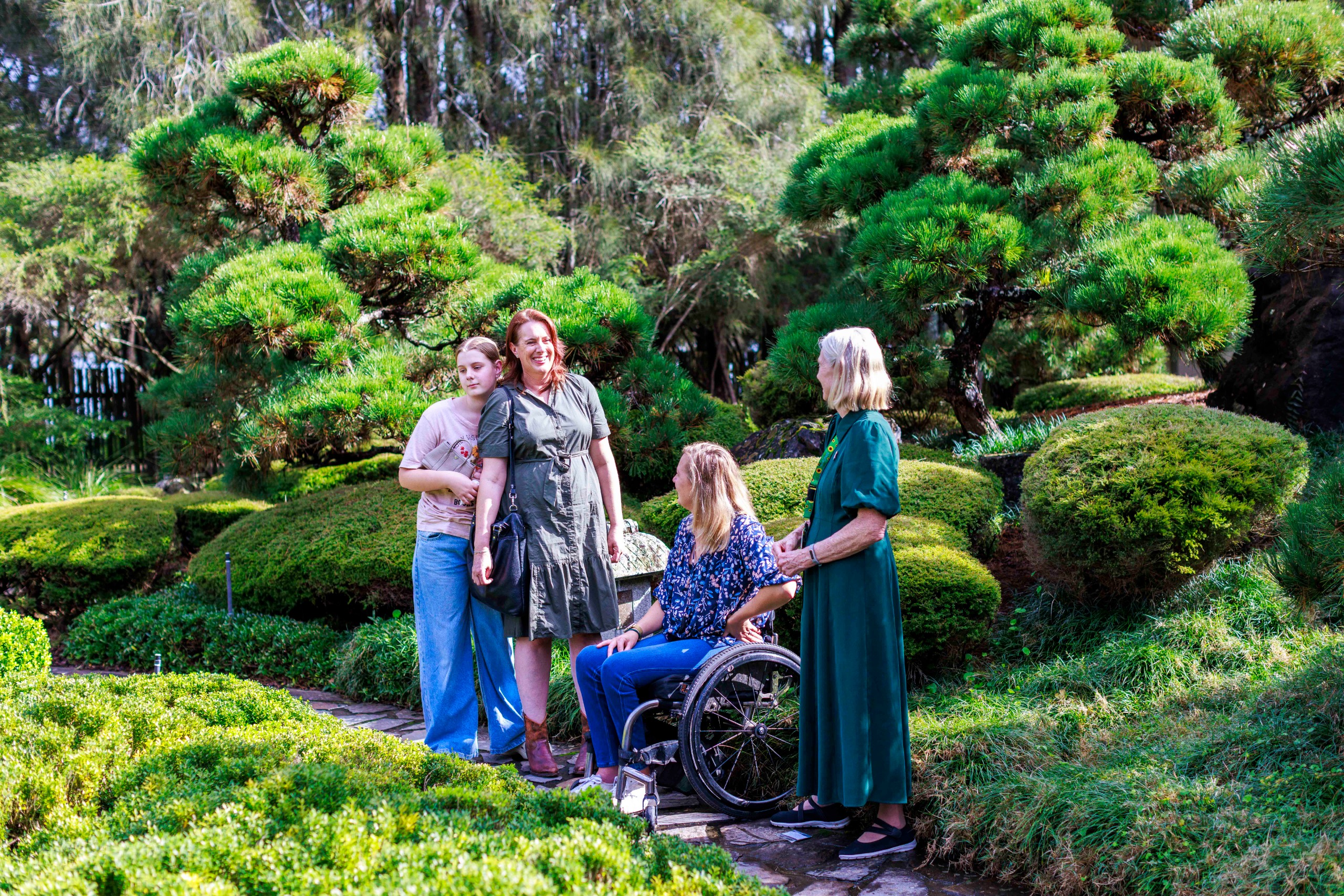 A woman in a wheelchair surrounded by three standing women in a vibrant garden with lush trees