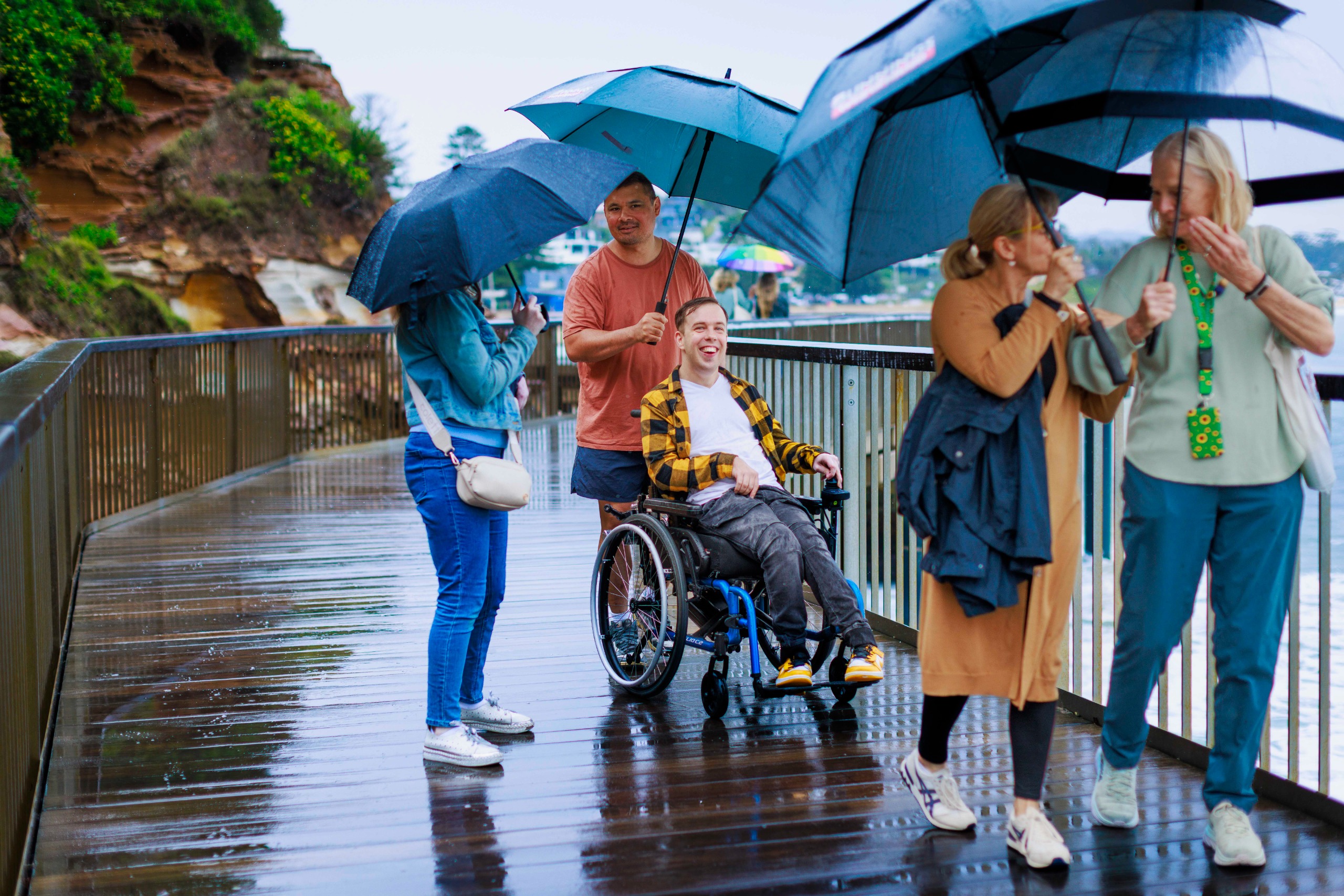 A group walks on a rainy boardwalk with umbrellas, including a man in a wheelchair and a woman with a hidden disabilities lanyard