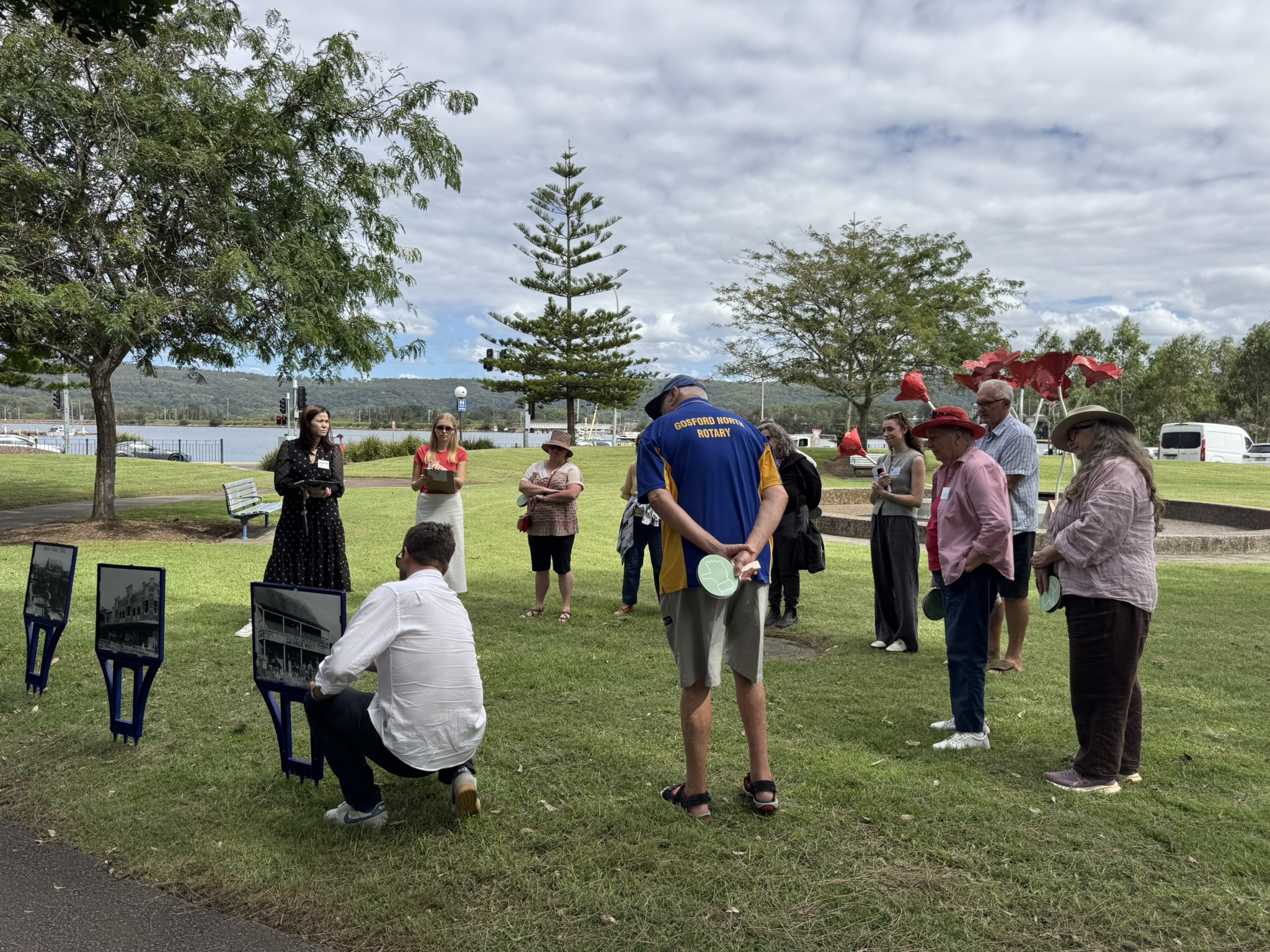 A group of people standing in a park, with one man talking to the group while crouched and pointing at a sign