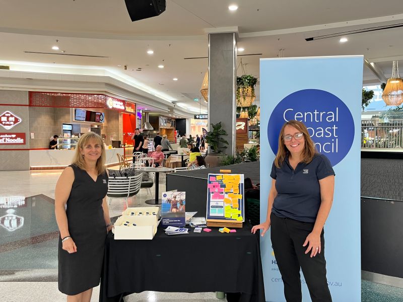 Two women are next to a table featuring a Central Coast Council sign during a community pop-up event.