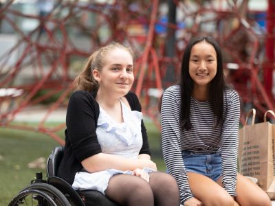 a girl in a wheelchair and a girl sitting on a bench smiling at the camera with a playground in the background